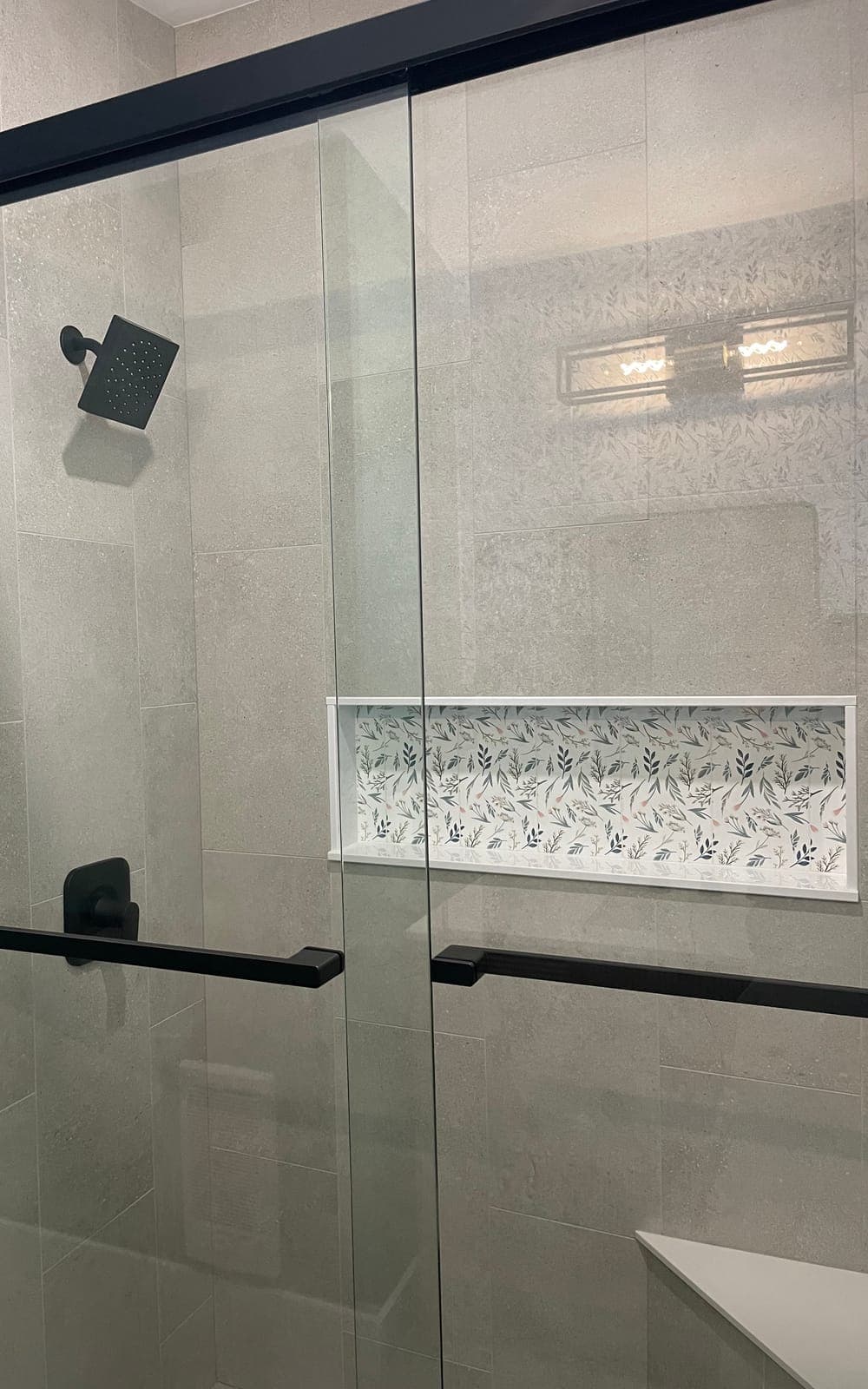 The same shower after: warm grey tile, a black shower head, black-framed glass doors and a leaf-pattern tile niche