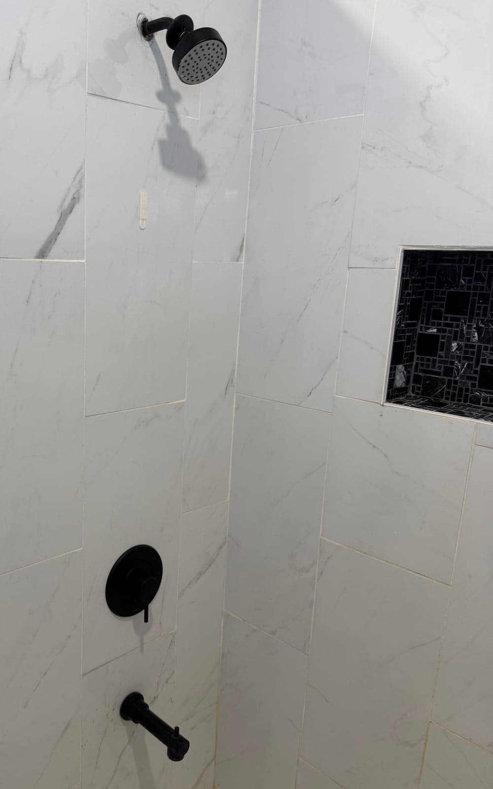 The shower before: white marble-look wall tile, a dark mosaic niche, and a shower head and valve
