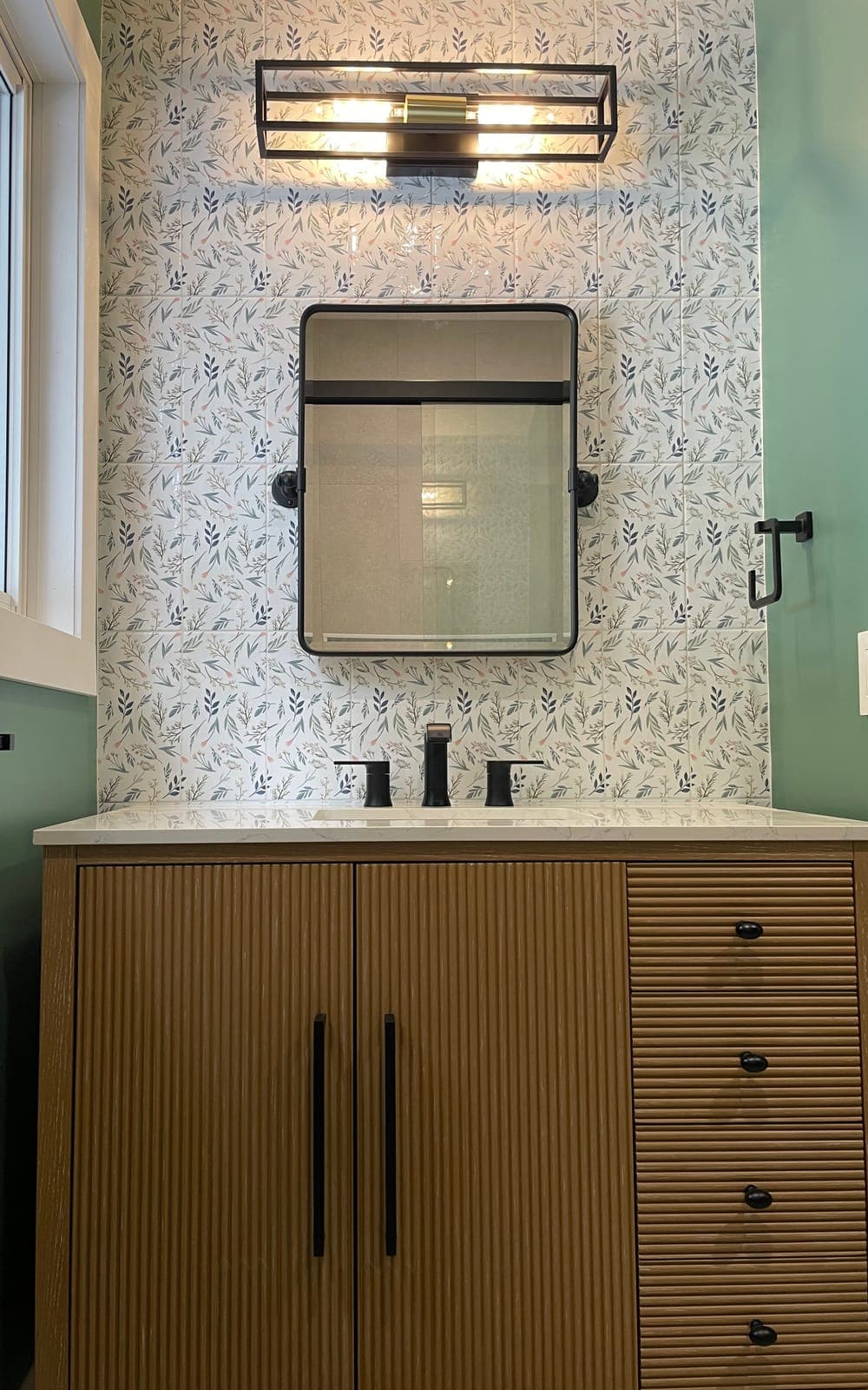 A new wood vanity with black hardware, under a mirror and light on a leaf-pattern feature tile wall