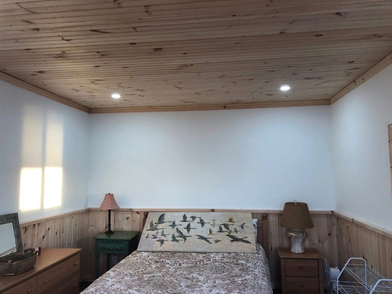 Inside the bunkie: a tongue-and-groove pine ceiling with crown trim and pot lights, white walls above pine wainscoting, and a bed