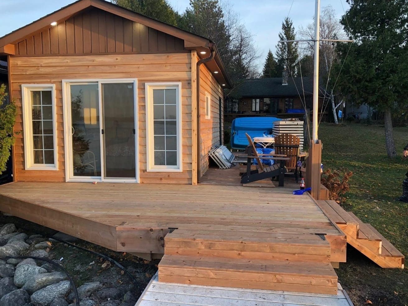 A waterfront bunkie with cedar lap siding, board-and-batten in the gable, pot lights in the soffit, a sliding patio door between two windows, and a wide new deck with broad steps down to the dock