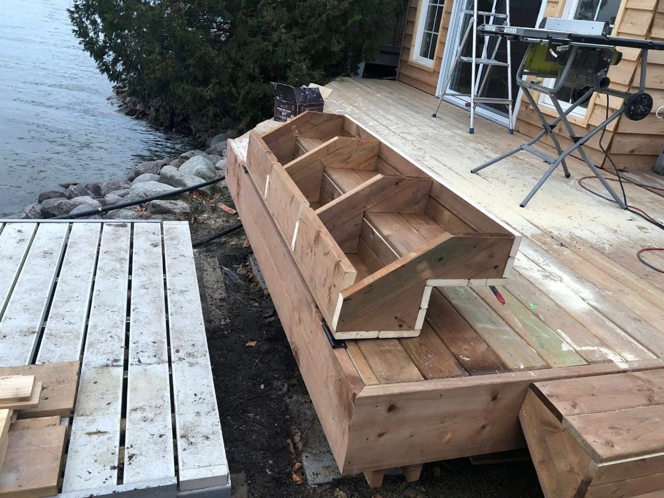 The bunkie deck mid-build beside the lake: wide steps being framed and boarded, with a table saw set up on the new decking