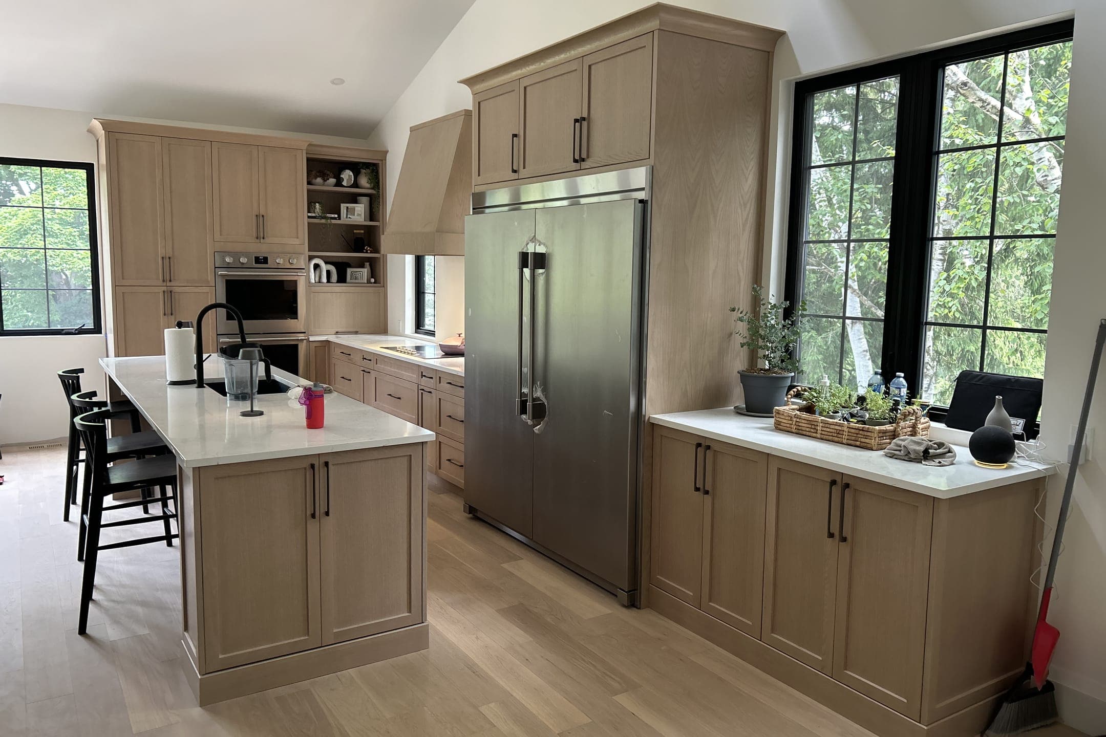 A kitchen with light wood shaker cabinets, a white-topped island, a built-in stainless fridge and black-framed windows under a vaulted ceiling