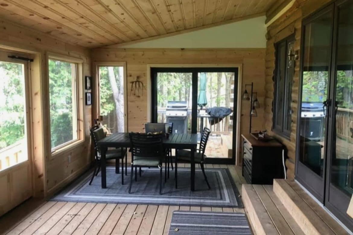 The same sunroom’s dining end: a pine ceiling, patio doors out to a deck, and a log wall with steps up to the house