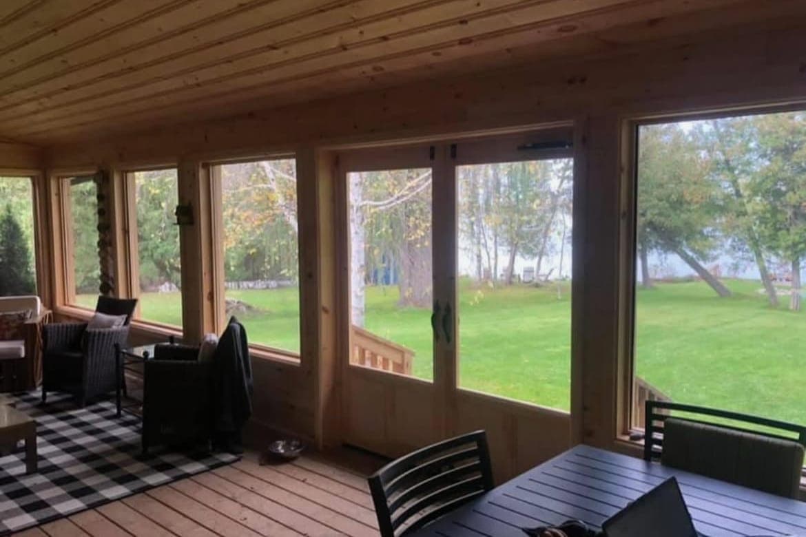 A three-season sunroom with pine walls and ceiling, and wide screened windows looking out to a lawn and trees