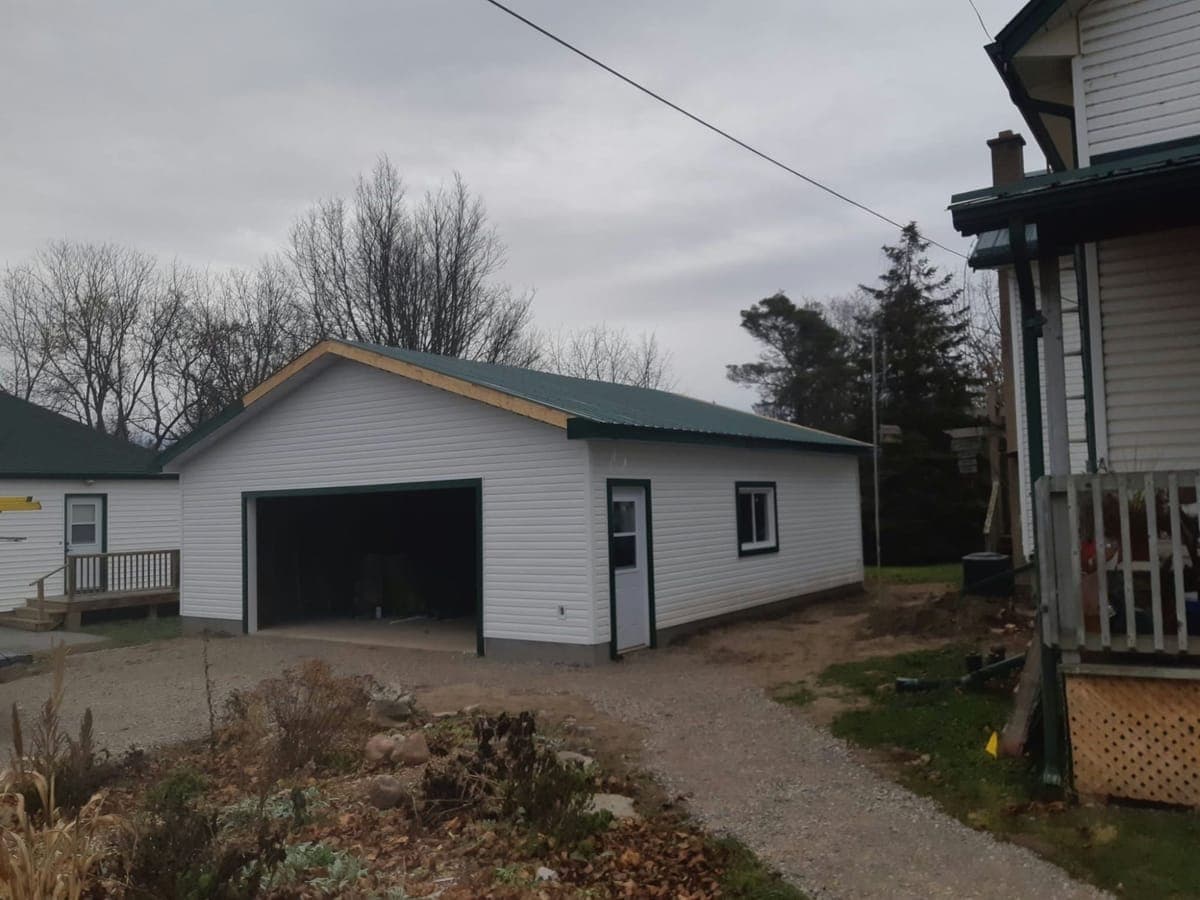 A finished double garage with a gable roof, white vinyl siding, a green steel roof and trim, a man door and a window on the side