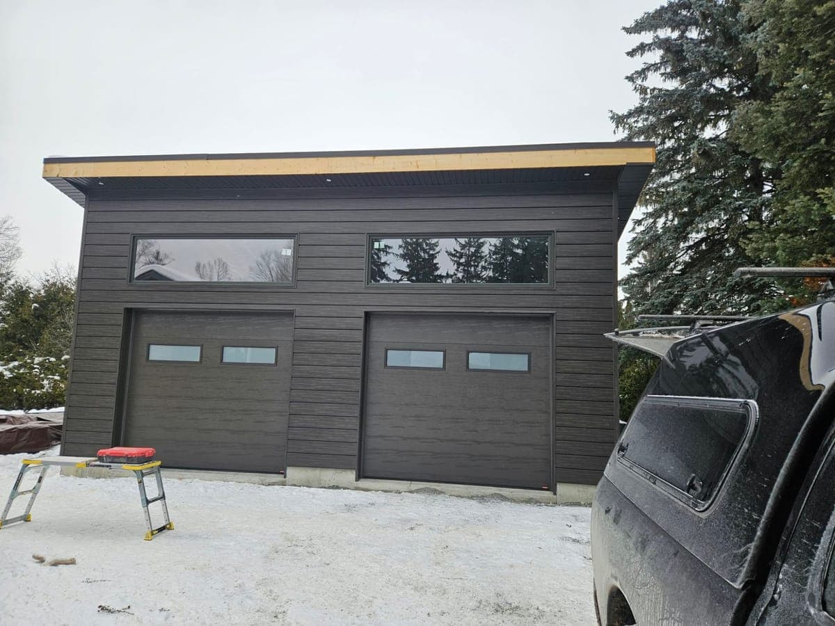 A new double garage with a flat roof, dark horizontal siding, two insulated overhead doors, long transom windows above them and pot lights in the soffit