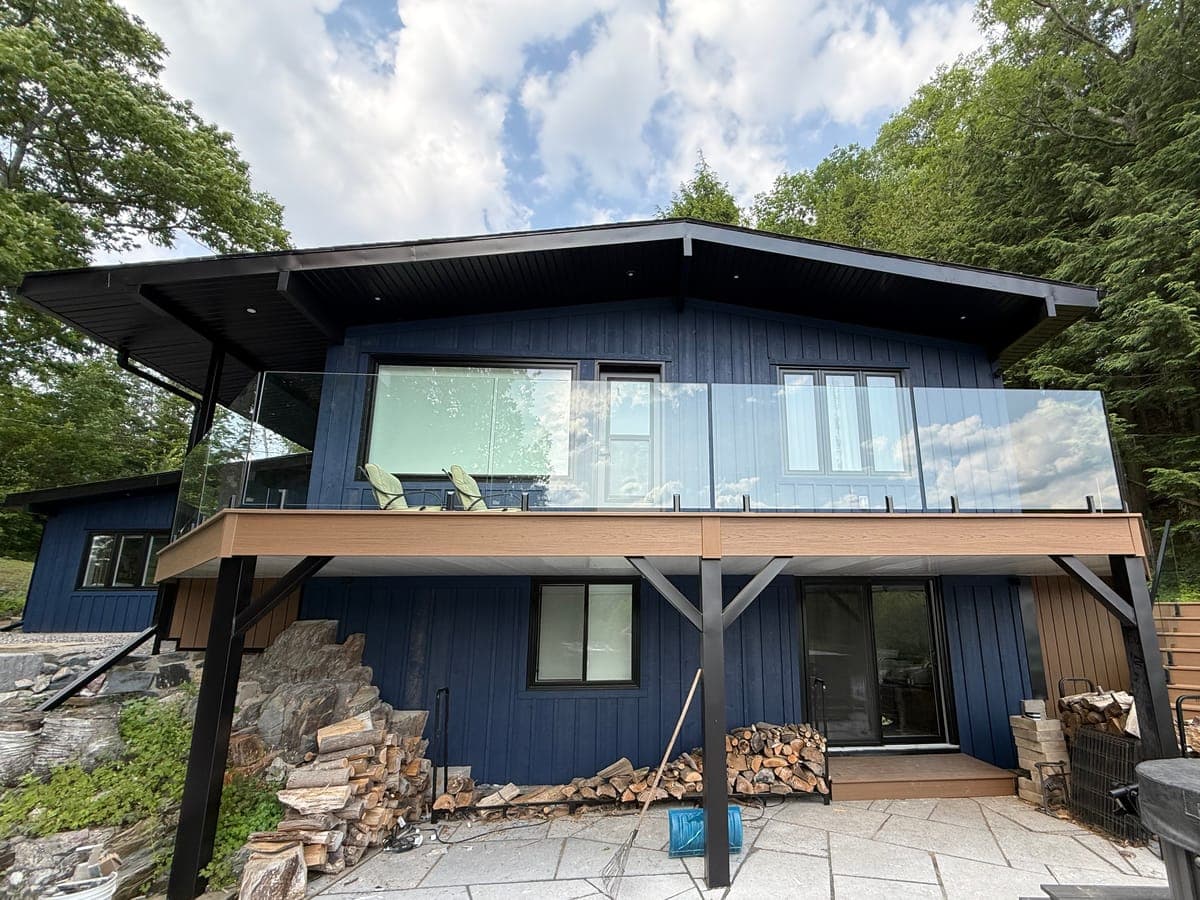 A waterfront cottage renovated by Mega Construction: a raised deck with glass railings, dark blue board-and-batten siding, and black windows and doors